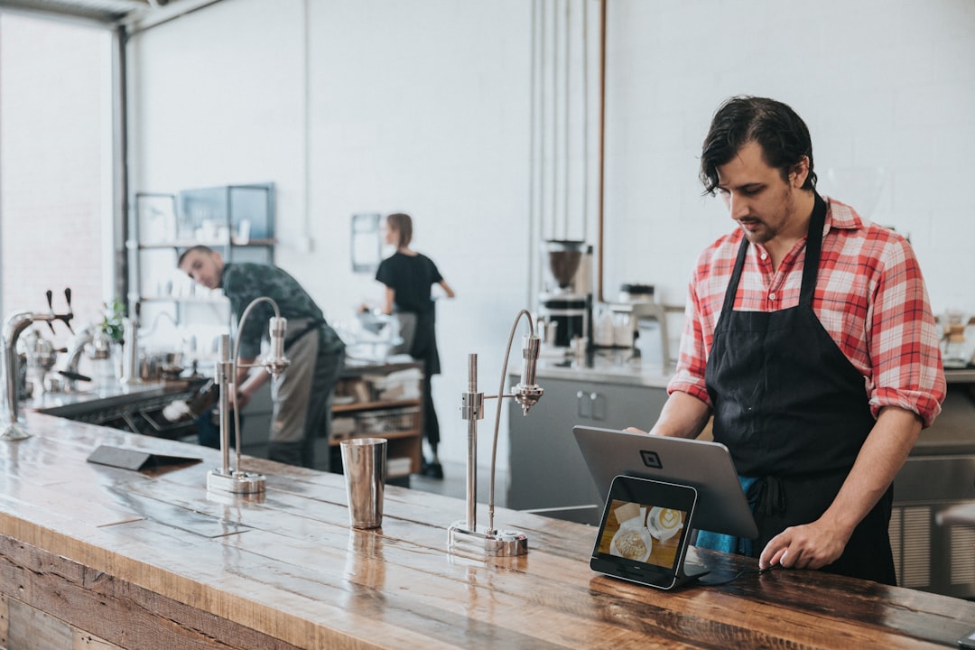 A barista in a plaid shirt and apron using a tablet at a wooden counter